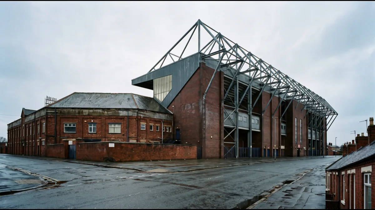 A tall modern brick and steel football stand joined to an older lower stand, seen from street level on an overcast day