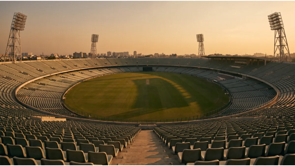 A vast empty oval stadium bowl seen from a high upper tier at golden hour, tiers of grey and green seats sweeping around a bright open playing surface