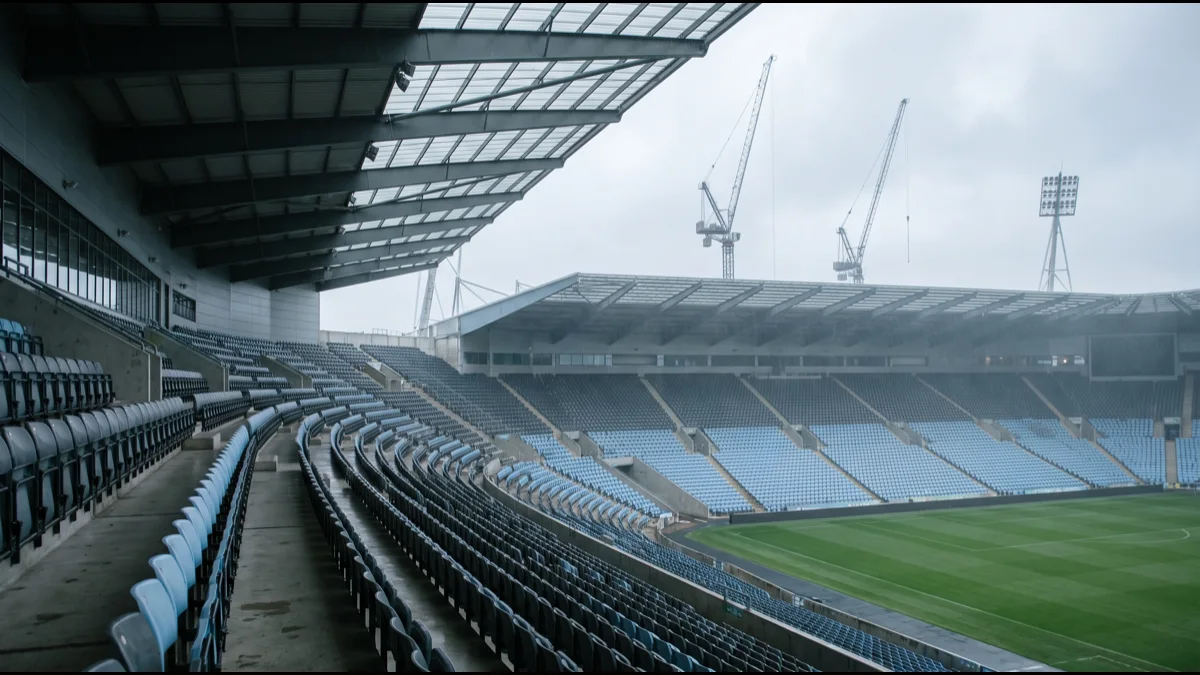 Empty tiered stadium seating curving away under a grey sky, with floodlight pylons and construction cranes visible beyond the roofline