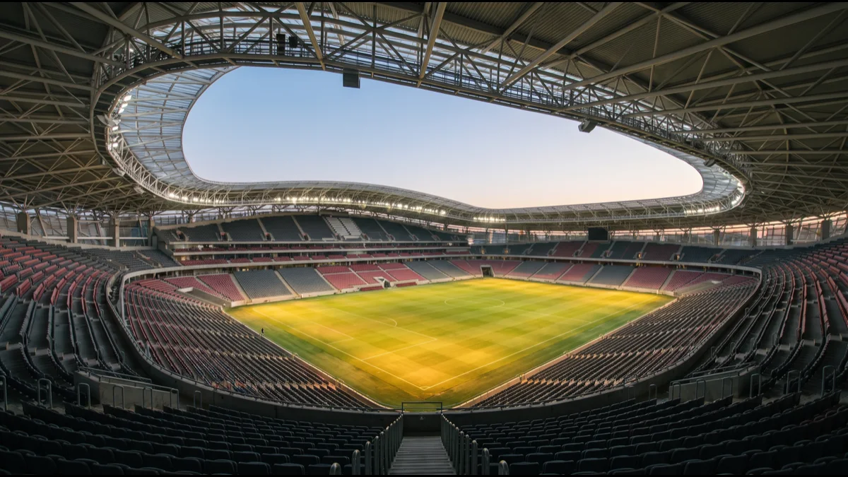 An empty modern stadium bowl at dusk with a lattice roof, floodlights on and the pitch bright below, seen from the upper tier