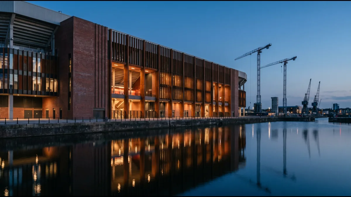 A newly built stadium exterior in dock-side surroundings at dusk, brick and steel facade lit from within, with cranes on the skyline behind