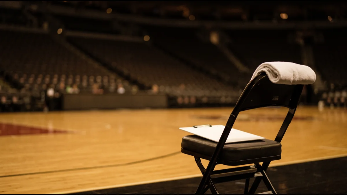 A plain folding courtside chair on a polished hardwood basketball court with a blank clipboard on the seat and a rolled towel over the back