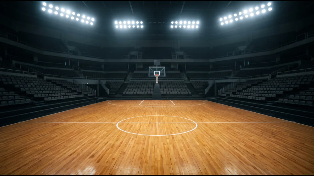 An empty indoor basketball arena with polished hardwood floor and tiered seating, lit by overhead spotlights from above the court