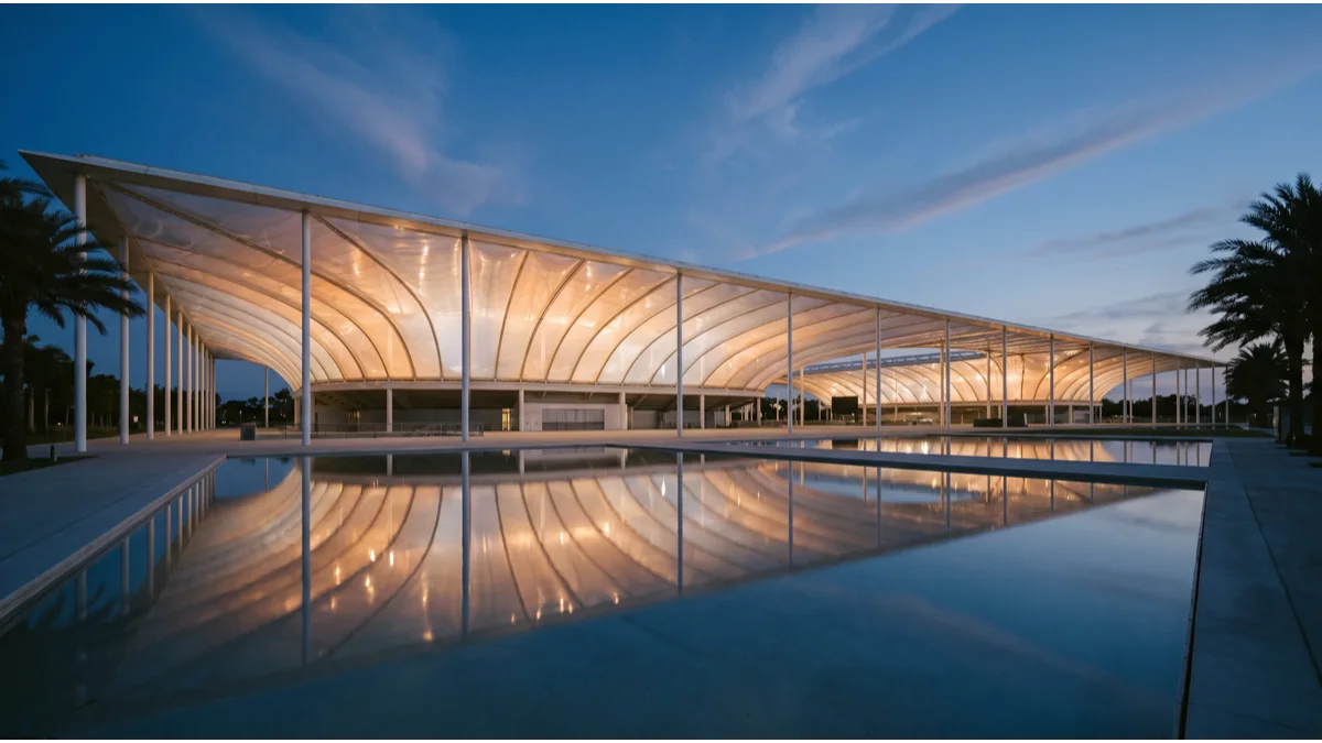 A vast translucent-roofed stadium exterior at dusk with a sweeping canopy and reflecting pool in the foreground, lit from within