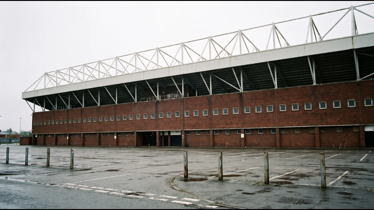 A large red-brick football stand with a cantilevered roof seen from outside on an overcast day, with an empty forecourt in front