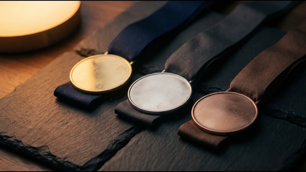 Three blank unmarked medals on ribbons laid on a dark slate surface, lit by a single warm lamp against deep shadow