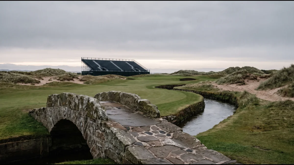 A links golf course at dawn with a stone road bridge over a burn in the foreground, dunes and a distant grandstand behind under grey sky