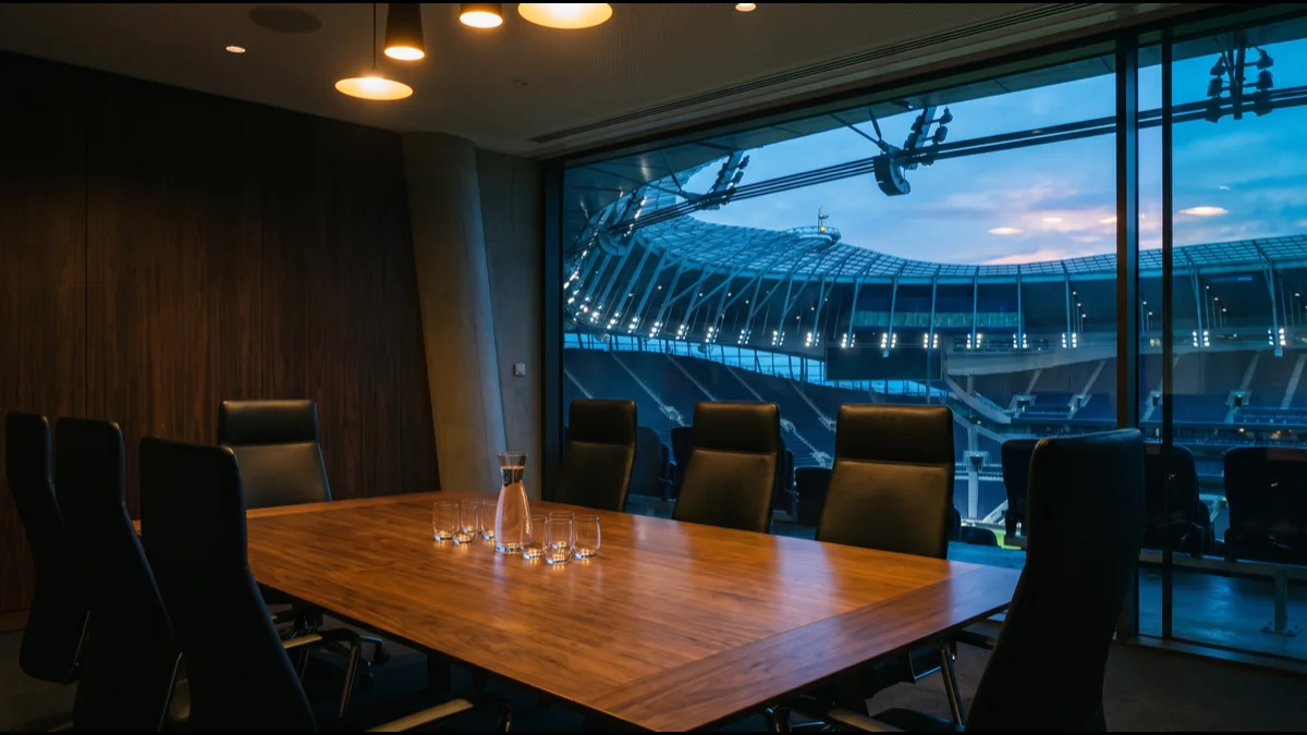 An empty boardroom table with leather chairs beside a tall window overlooking a floodlit football stadium at dusk