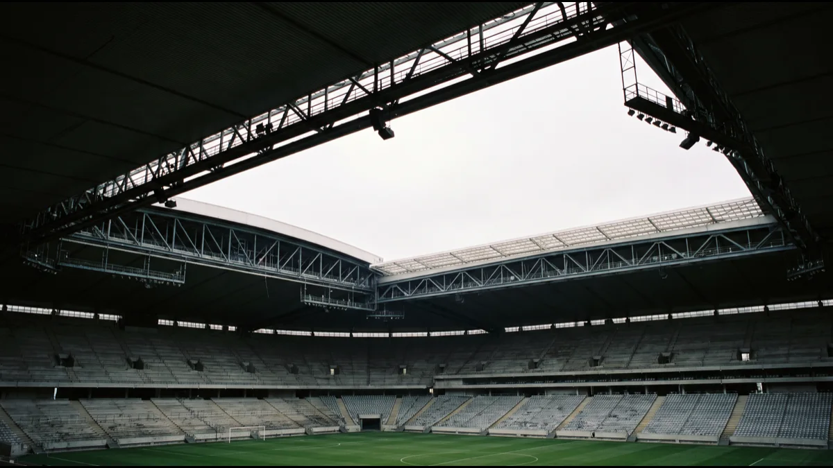 The interior of a large empty stadium seen from the lower tier, its retractable roof half open with a bright slot of overcast sky above rows of grey seats