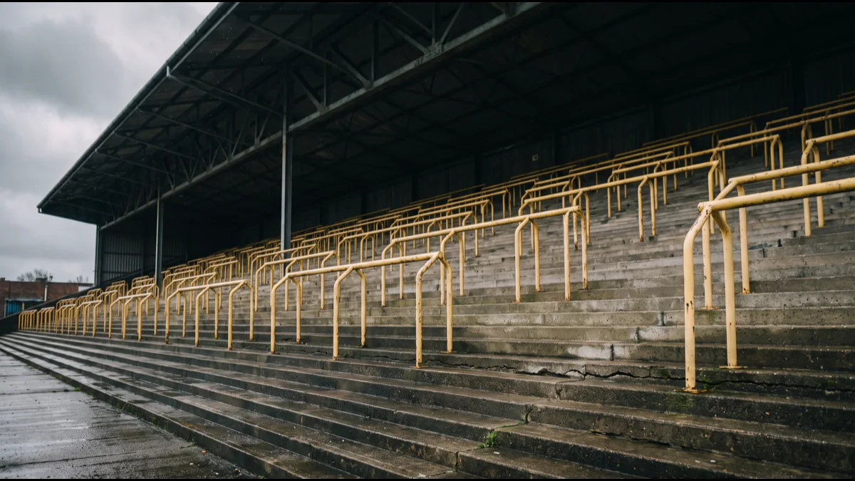 A vast empty single-tier standing terrace in a football stadium, rows of yellow crush barriers receding steeply upward under a dark steel roof