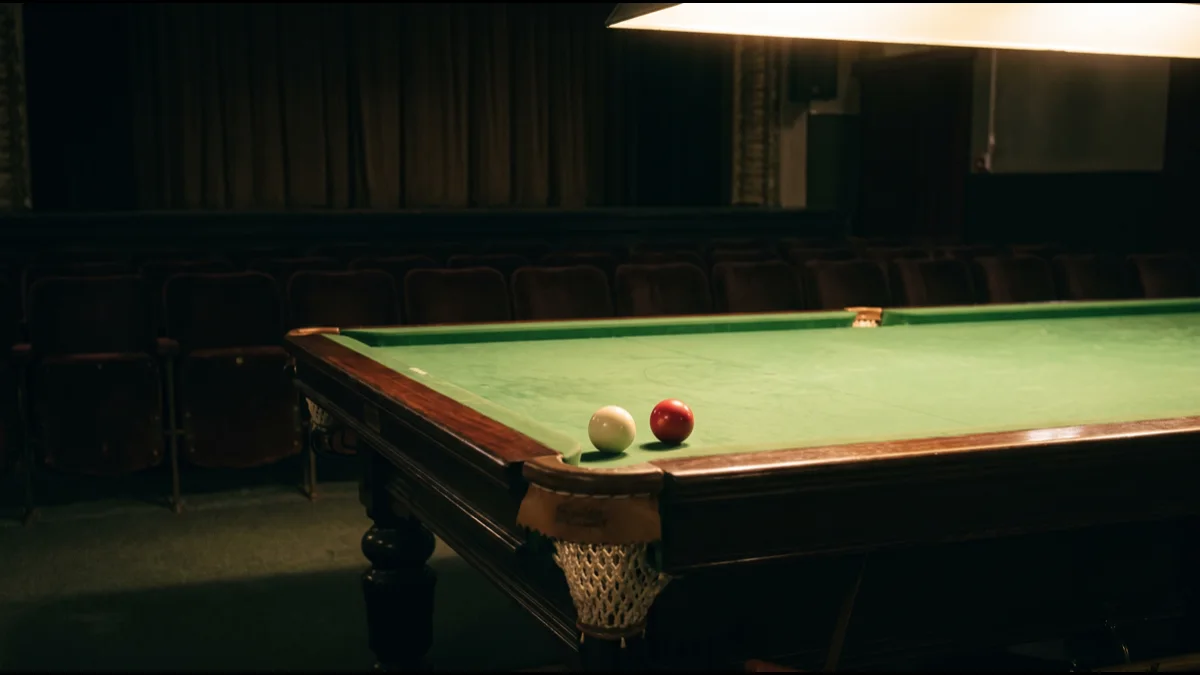 A white cue ball and a single red ball resting on green snooker baize beside a corner pocket, lit from above, with rows of empty dark auditorium seats behind