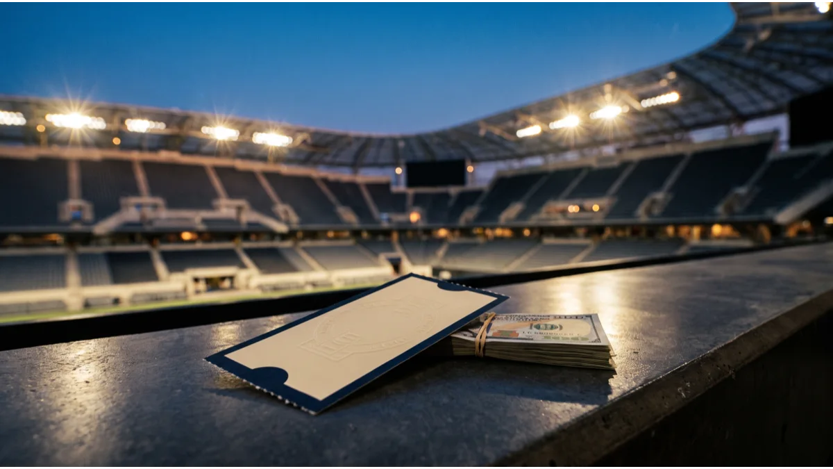 An embossed event ticket stub and a banded stack of banknotes on a concrete ledge, with a floodlit stadium bowl blurred behind at dusk