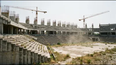 A half-built concrete sports venue with exposed rebar and idle cranes under a bleached sky, weeds growing through the site
