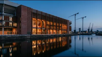 A newly built stadium exterior in dock-side surroundings at dusk, brick and steel facade lit from within, with cranes on the skyline behind