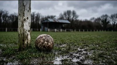 A muddy leather football in wet grass beside a peeling white corner post on a rough pitch, a small tin-roofed terrace out of focus behind