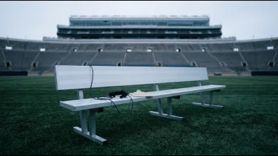 An empty aluminium coach's sideline bench on stadium turf at dawn with a coiled headset cable and a blank clipboard, empty tiers behind