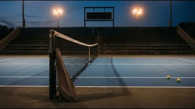 An empty floodlit tennis court at dusk with a racket bag resting against the net post and a scoreboard switched off