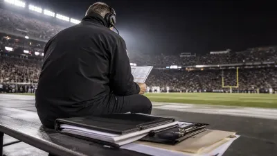Anonymous football head coach wearing a headset and studying a play sheet under stadium lights