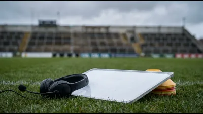 A plain black coaching headset and a blank white tactics whiteboard lying on wet grass beside training cones, empty stadium seating out of focus behind