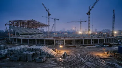 A large stadium under construction at dusk, a partly completed concrete bowl with exposed steel roof trusses and tower cranes standing over it