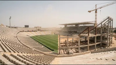 A vast empty stadium bowl under scaffolding with one stand partially demolished, seen from the upper tier against a pale Mediterranean sky