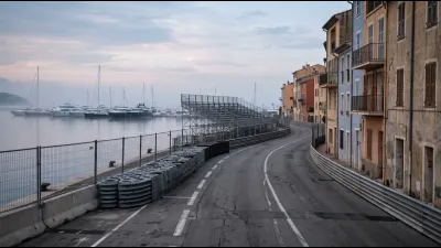 An empty Mediterranean harbour street circuit at dawn, steel debris fencing and stacked tyre barriers lining a narrow road, moored yachts out of focus beyond