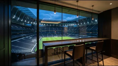A glass-walled executive suite high inside a large stadium, looking down over an empty bowl at dusk, with an empty desk in the foreground