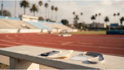 A phone face-down beside a printed session schedule and a plain accreditation wallet on a sunlit concrete bench, with a running track and empty stand blurred behind