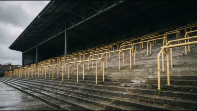 A vast empty single-tier standing terrace in a football stadium, rows of yellow crush barriers receding steeply upward under a dark steel roof