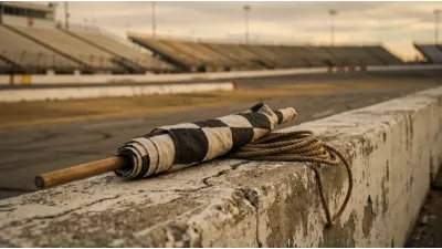 A furled black-and-white chequered flag resting on a weathered concrete pit wall, the banked oval and empty grandstand of a speedway out of focus behind