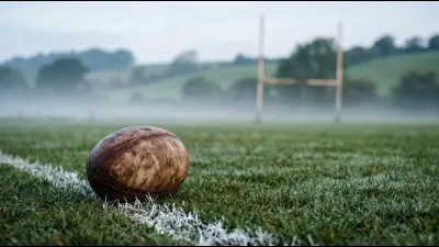A scuffed leather rugby ball on dewy grass beside a painted white touchline at dawn, mist across the turf and wooden goalposts out of focus behind