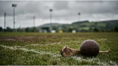 A worn leather ball and a plain ash stick with a curved blade resting on damp grass beside a painted white boundary line, a rural playing field behind