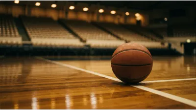 A plain unmarked basketball resting on a polished hardwood court beside a painted white line, the empty tiered seating of an arena out of focus behind
