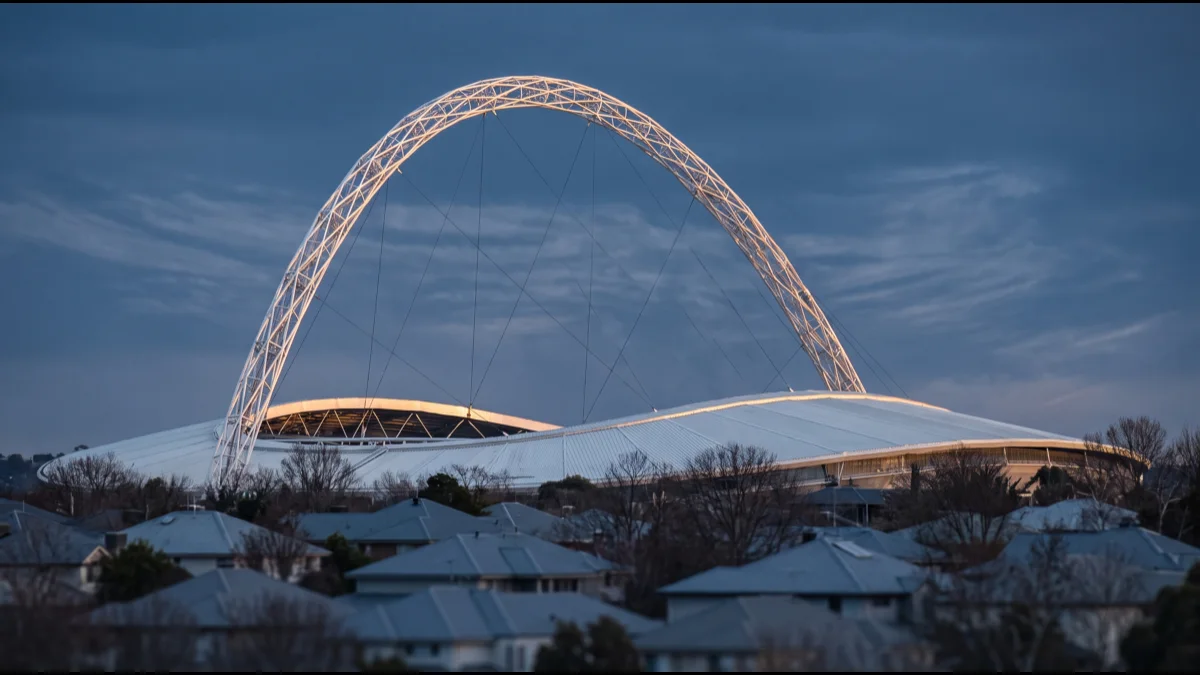 A tall white steel arch rising above a modern stadium roof at dusk, seen from a distance across suburban rooftops