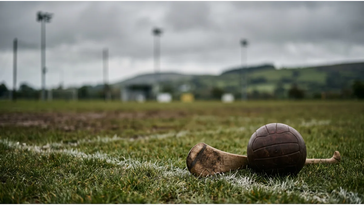 A worn leather ball and a plain ash stick with a curved blade resting on damp grass beside a painted white boundary line, a rural playing field behind