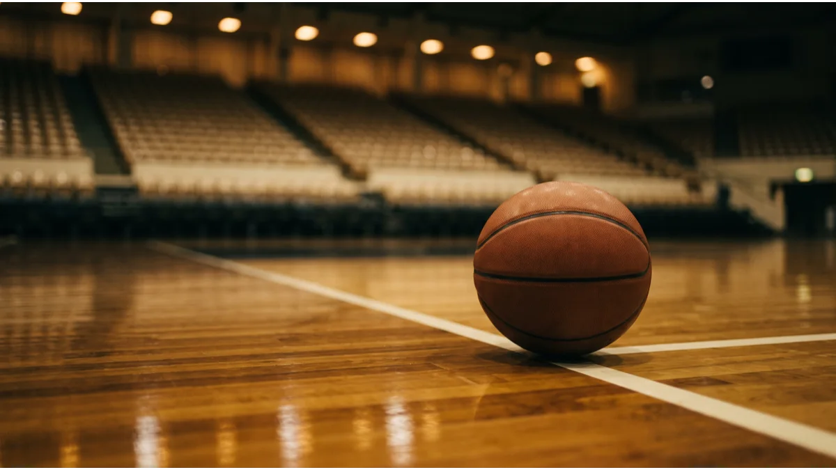 A plain unmarked basketball resting on a polished hardwood court beside a painted white line, the empty tiered seating of an arena out of focus behind